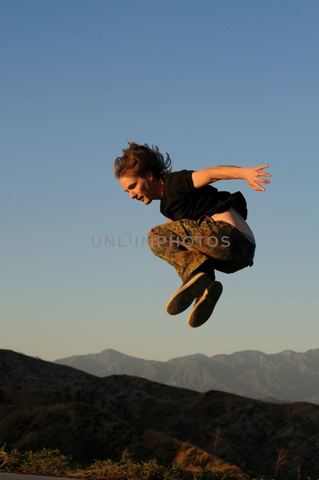 Caucasian male 20-40 in sportswear jumping above mountain peaks, face in profile to the left, one arm back, knees up.