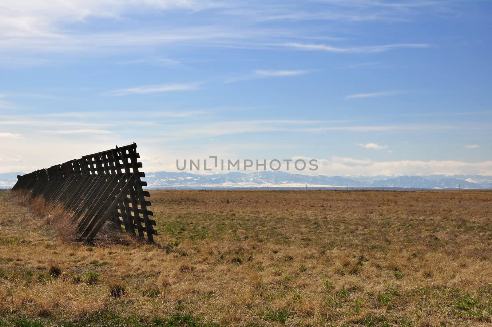 A snow fence stands in a field with the Rocky Mountains in the distance.