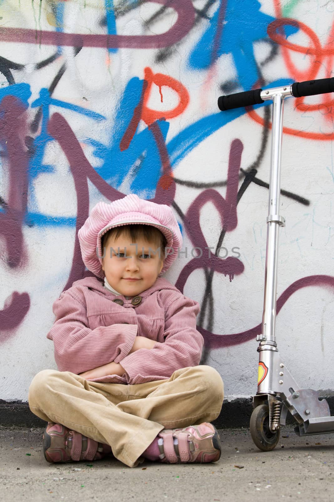 Funny little girl with scooter is sitting on the ground near graffiti painted wall