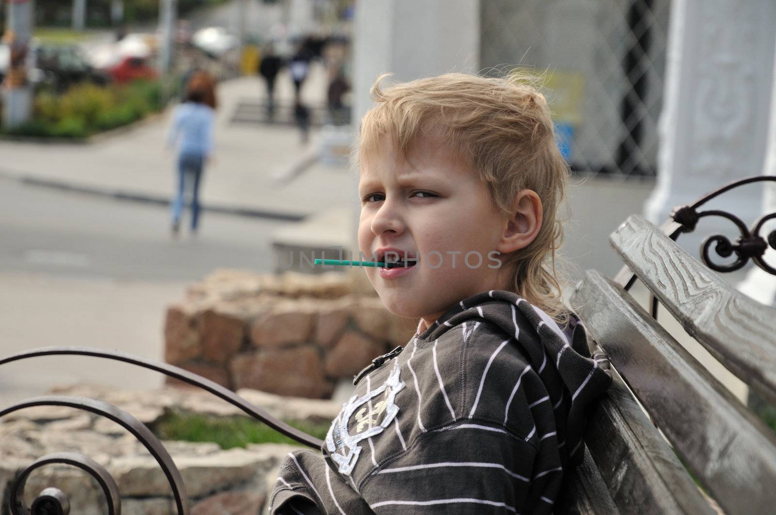 little boy is sitting on the bench and eating lollipop