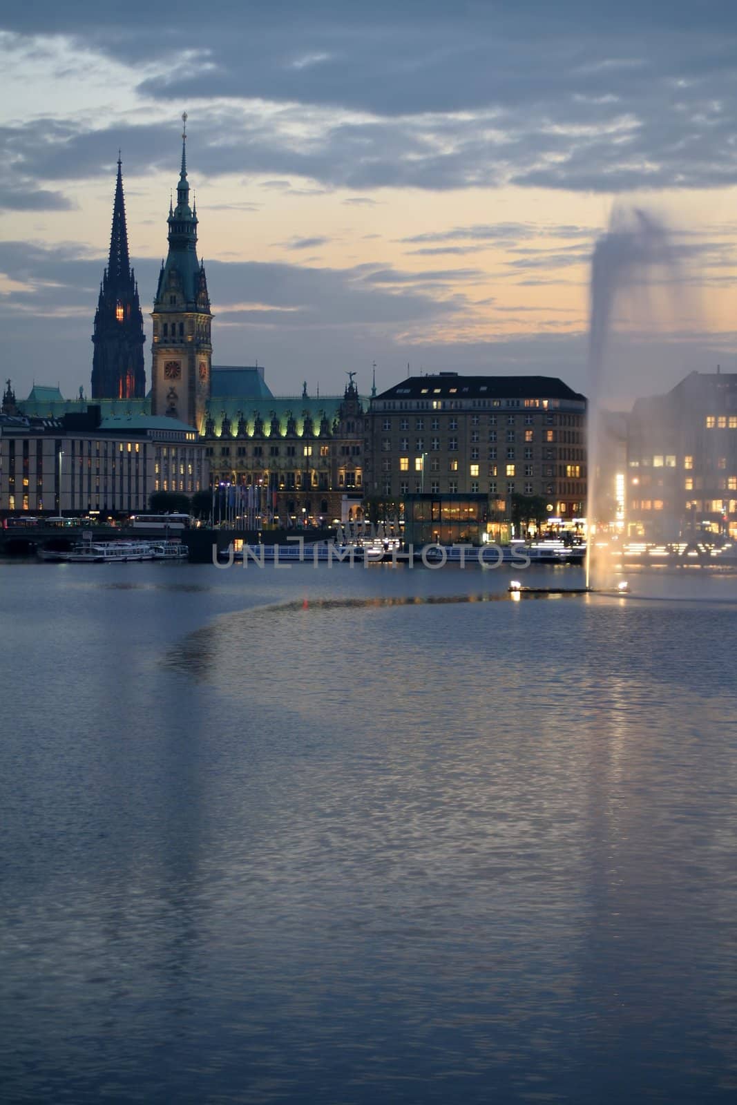 View across Hamburg's Alster lake at dusk, with illuminated water fountain. The skyline shows parts of the town hall and the Nikolai church.