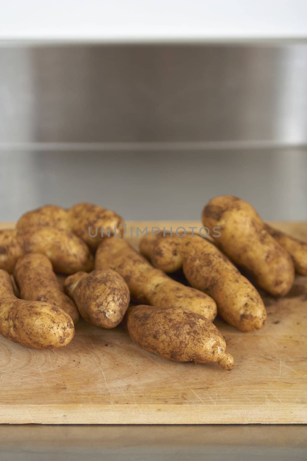 Wooden chopping board with fresh, dirty potatoes. Stainless steel bench in background. Shallow focus