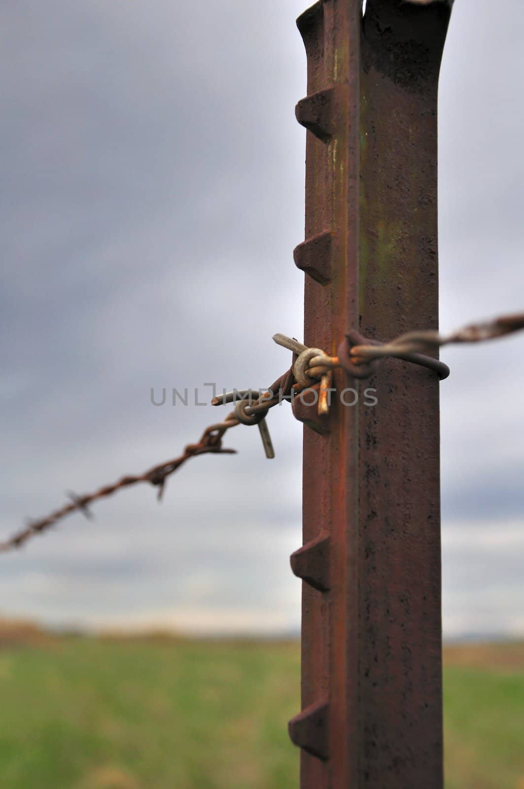 A rusty metal fencepost stands with an empty field blurred out in the distance.  