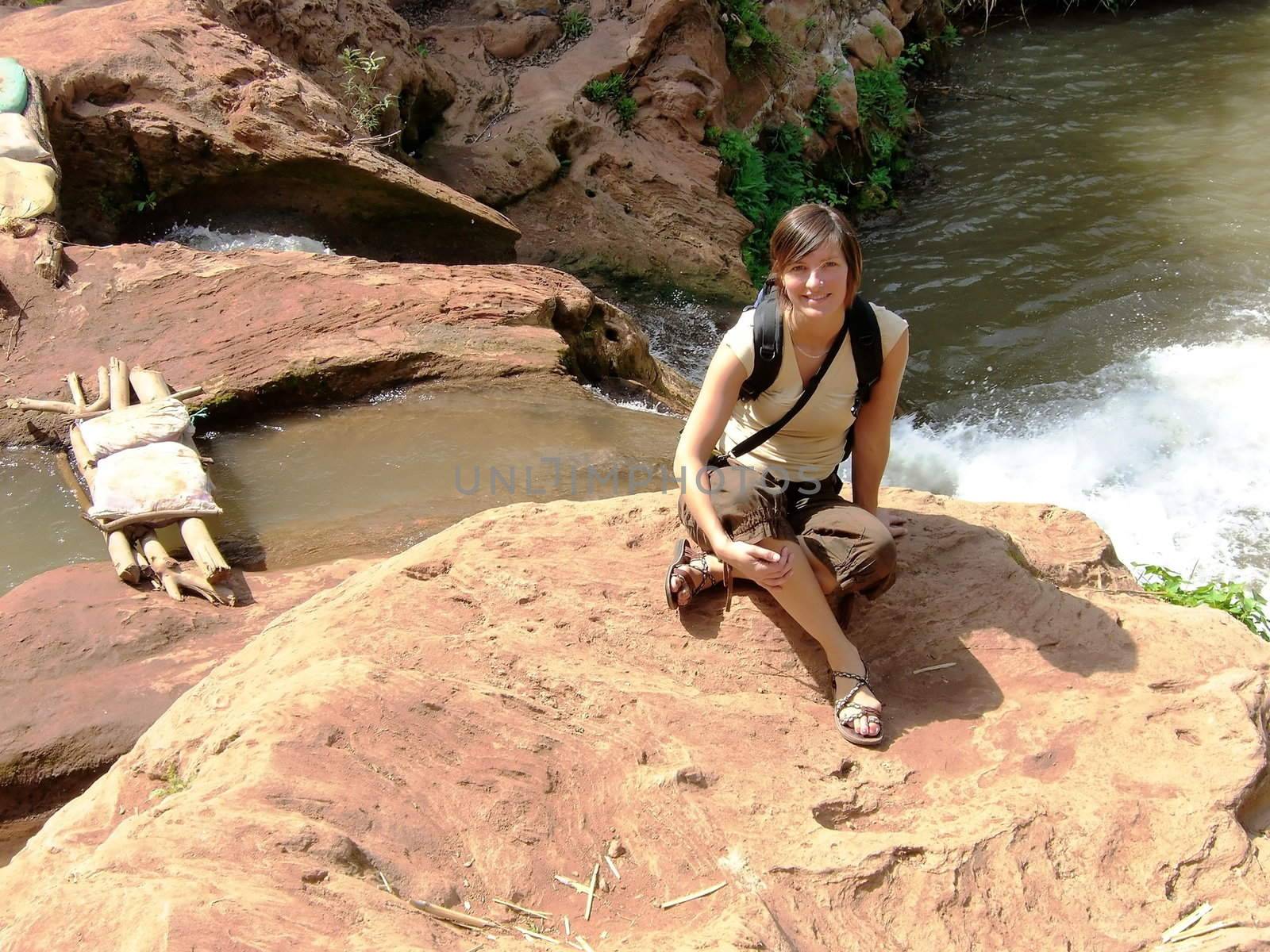 a beautiful woman is sitting on a rock beside a waterfall in Morocco