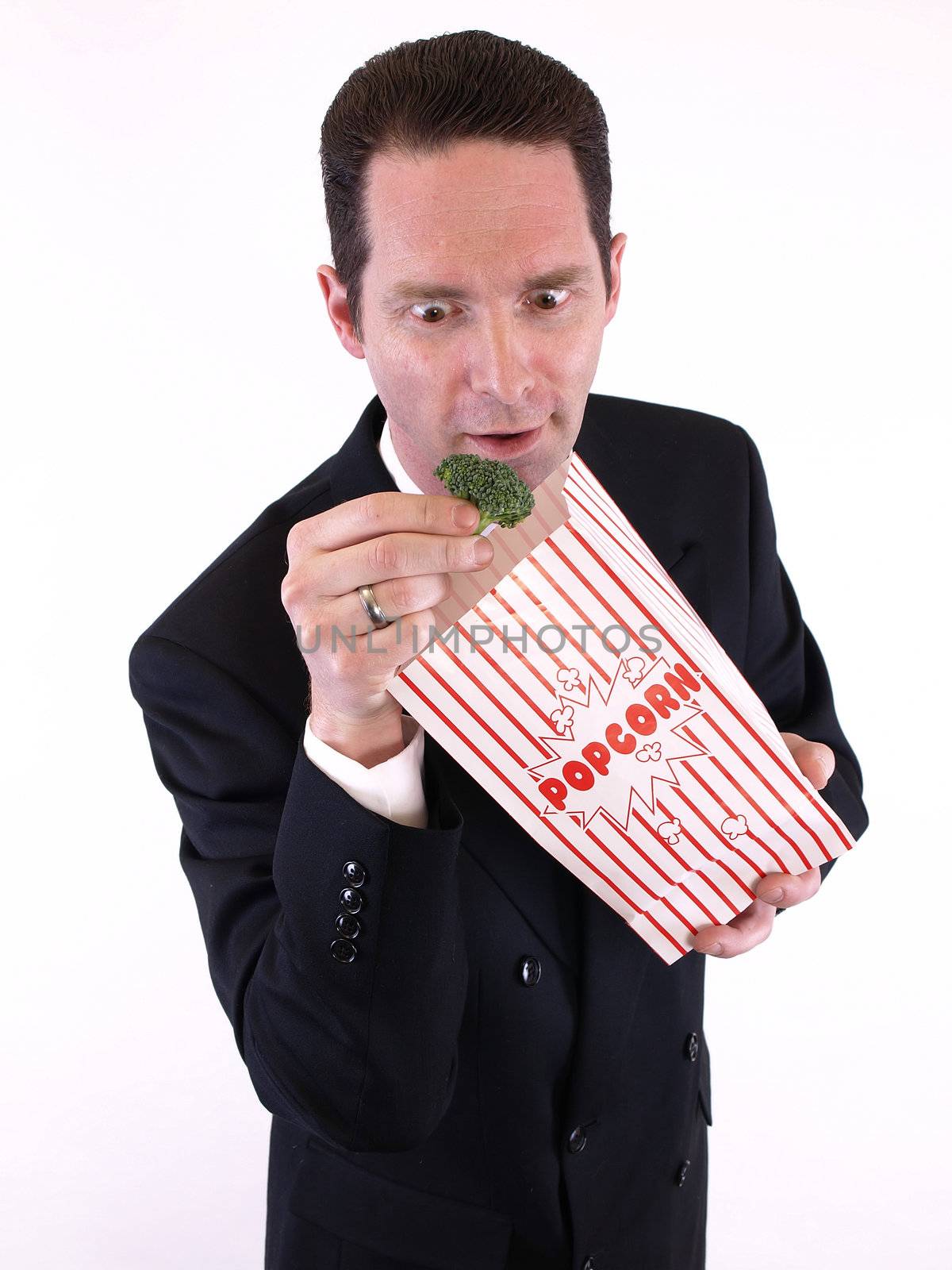 A man in a black suit pulls a piece of broccoli from a popcorn bag. Over white.