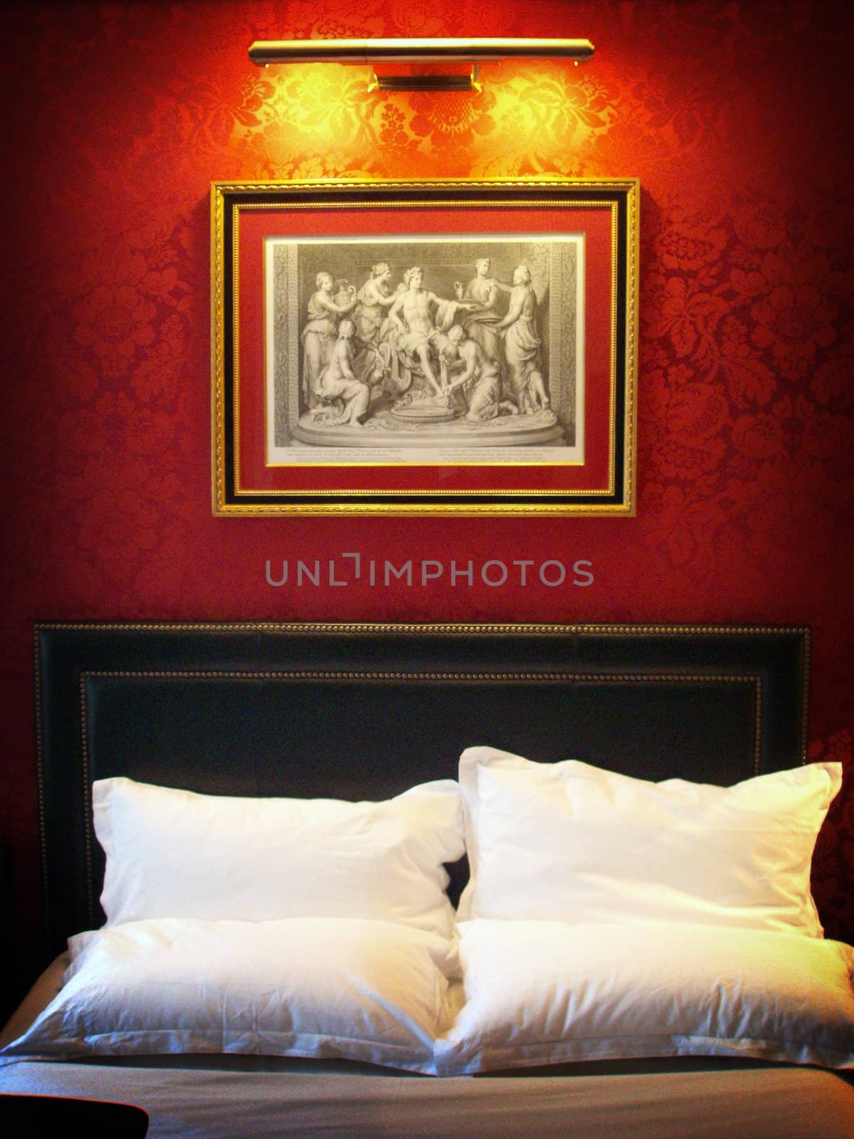Red wallpaper with art hanging on the wall above the top end of a bed in a Paris Hotel room.