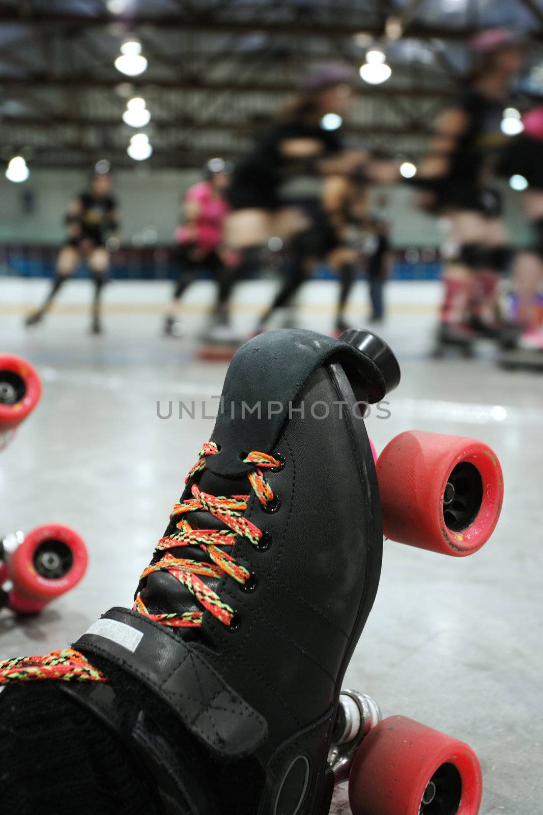 An abstract image of the roller-skates of a fallen skater as her teammates in the background continue to skate around the track of the roller derby.
