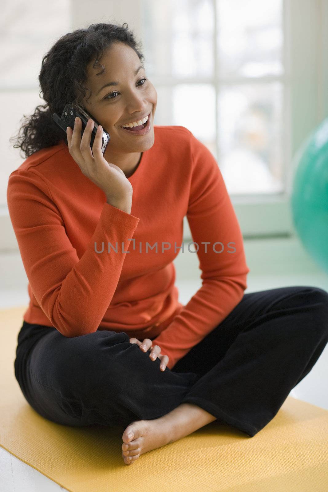 Smiling beautiful African American woman sitting on yoga mat talking on cell phone
