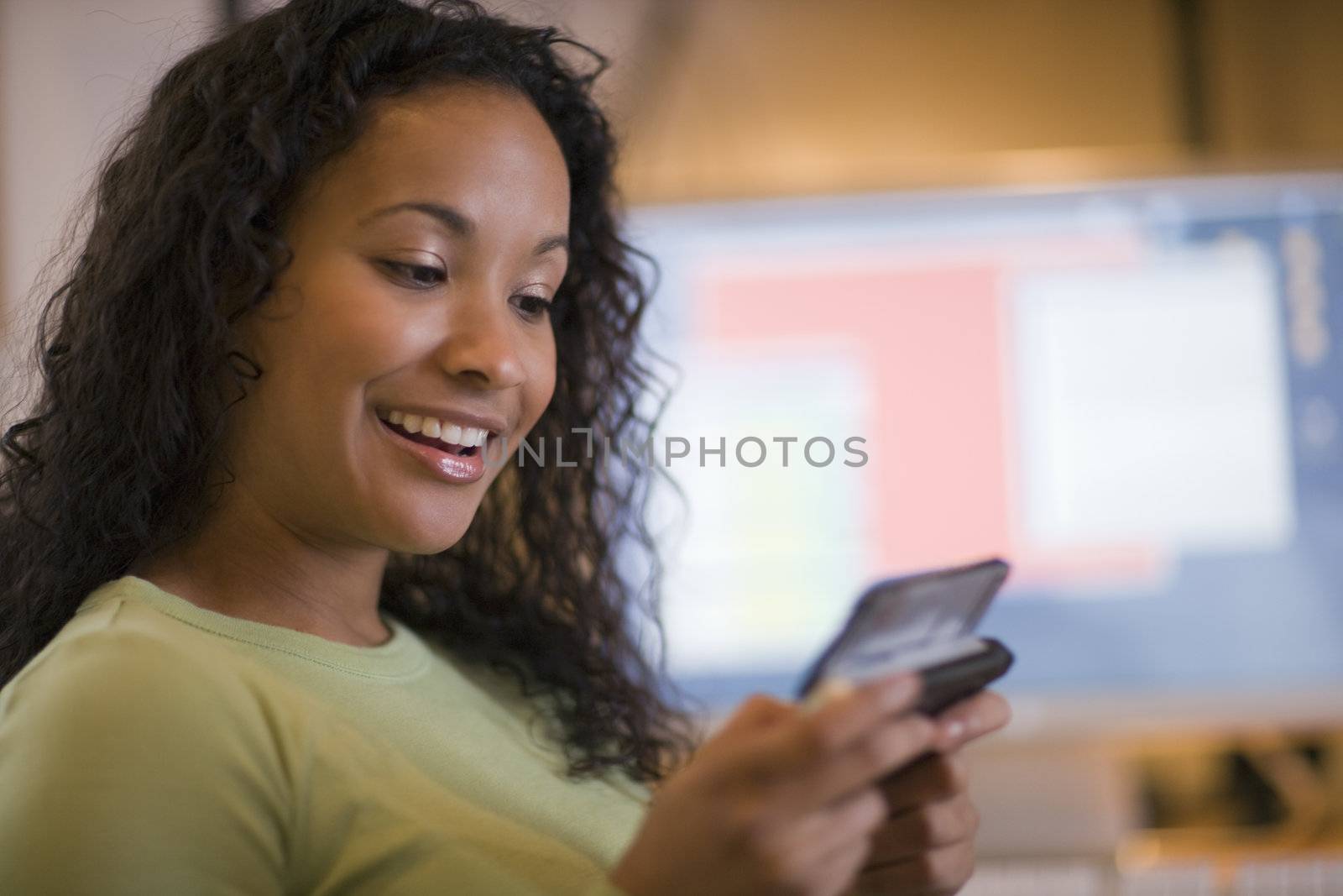Smiling young African American woman texting on cell phone with computer monitor in the background on desk
