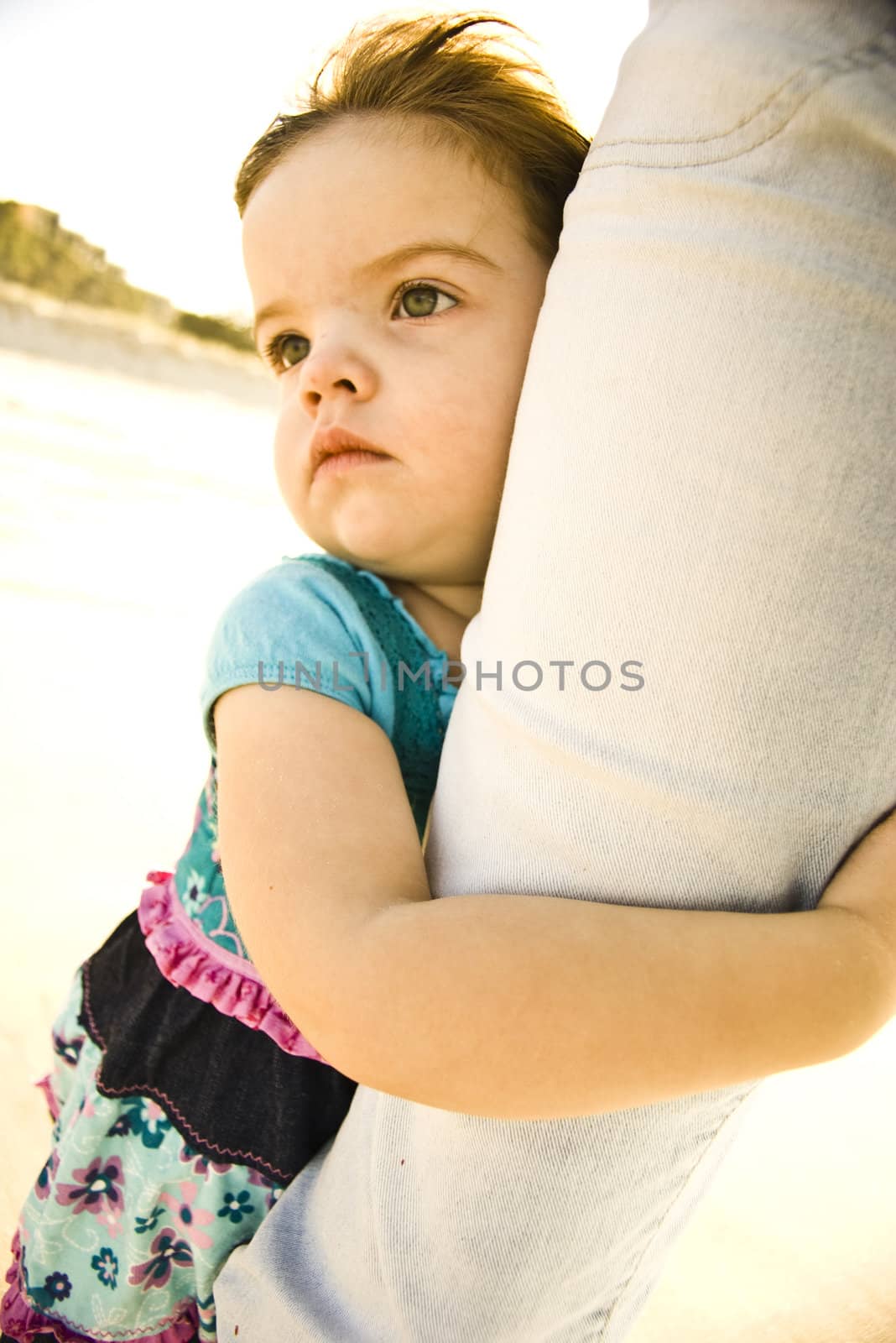 A young girl is clinging to her mem's leg at the  beach, facing the wind