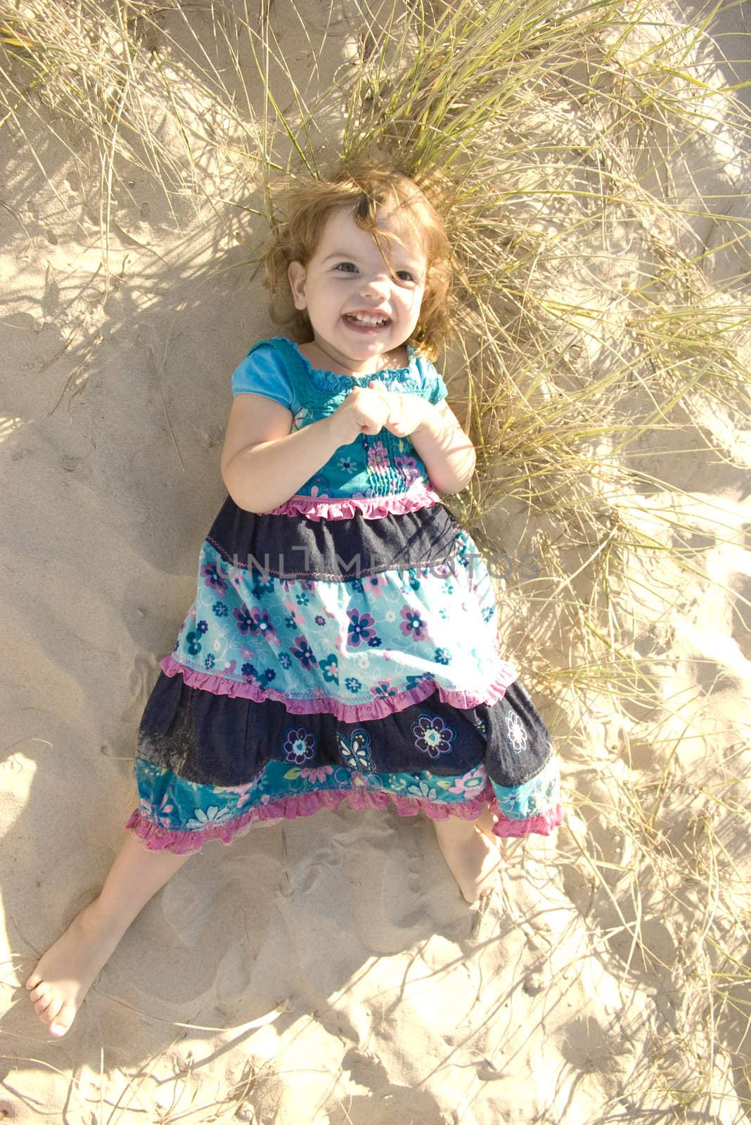 A little girl is laying on a sand dune in the sun, smiling