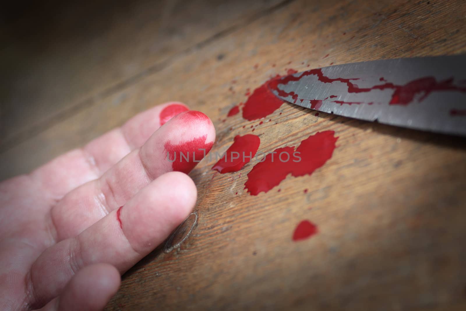 Low angled up close image of a bloodied hand and knife with blood splatters set on a wooden floor background.
