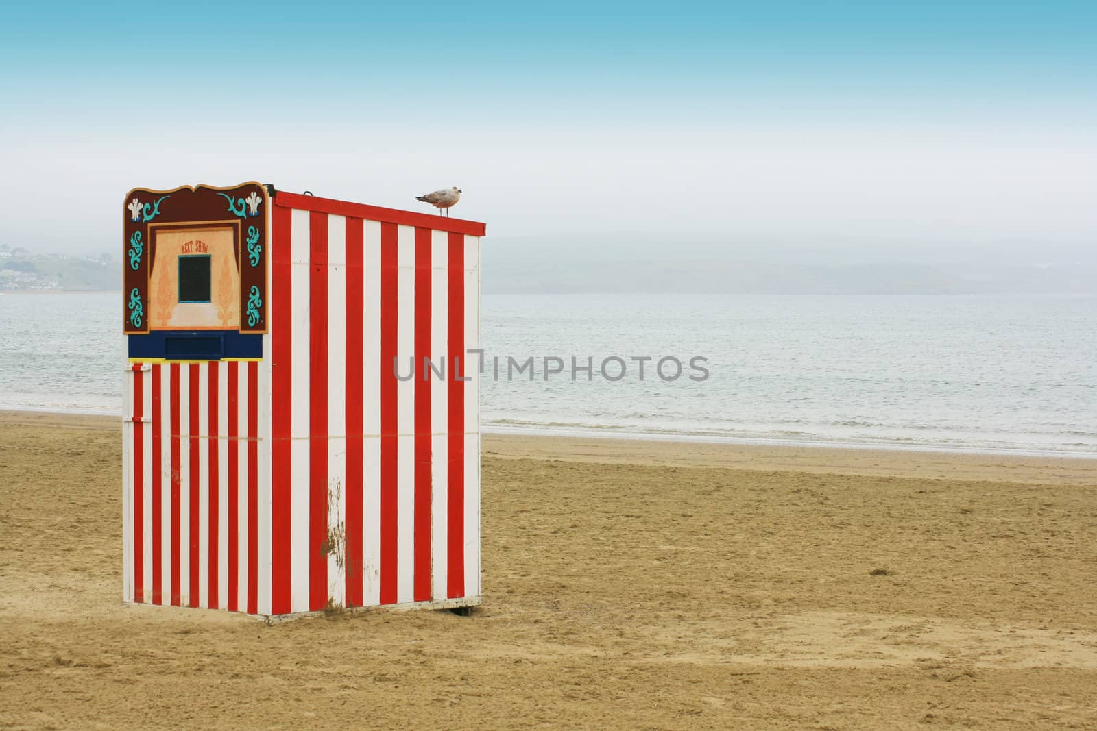 A red and white striped wooden punch and Judy booth on a beach located in Weymouth, Dorset UK. 
A seagull perches on top. landscape format, room for copy space etc.