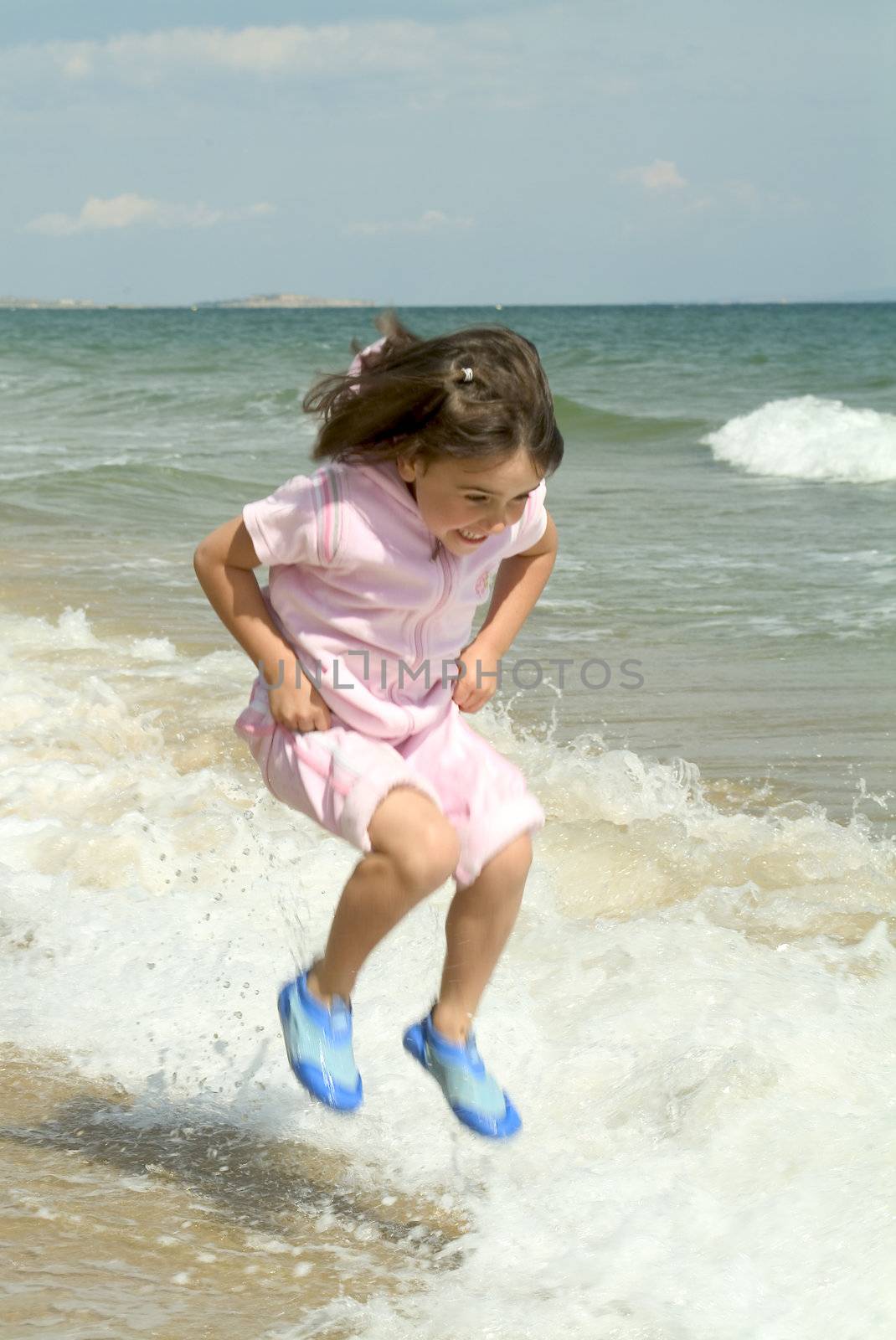 A LITTLE GIRL JUMPING OVER WAVES WHILE EXPRESSING EXCITEMENT AND JOY AT BEING IN THE WARMTH OF SUMMER ON THE BEACH BY THE SEASIDE AVOIDING GETTING WET AND GETTING SOME EXERCISE WHILE HAVING FUN AT THE EDGE OF THE SEA  