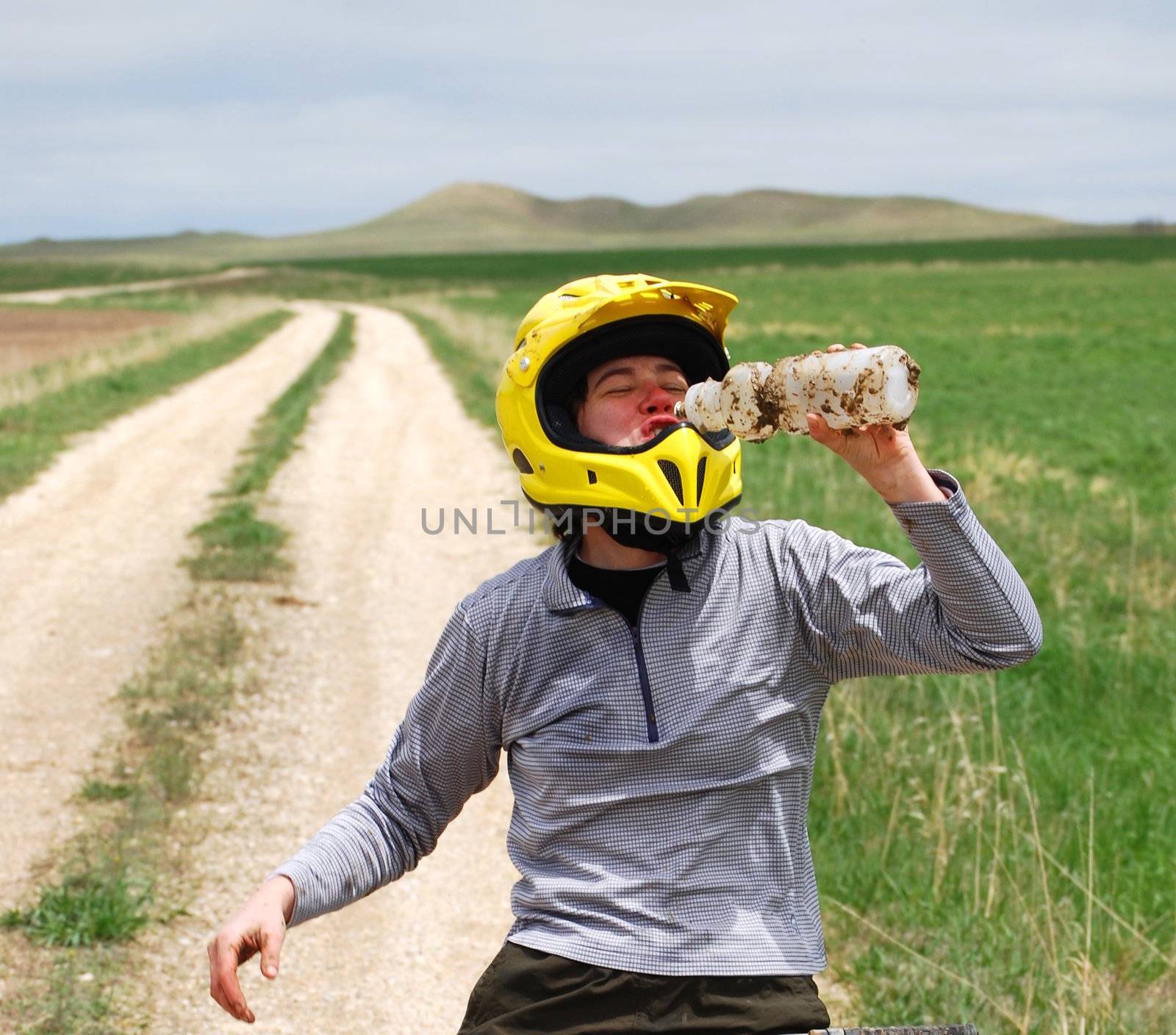 Mud-splattered mountain biker drinking water against a dirt road and green pasture.