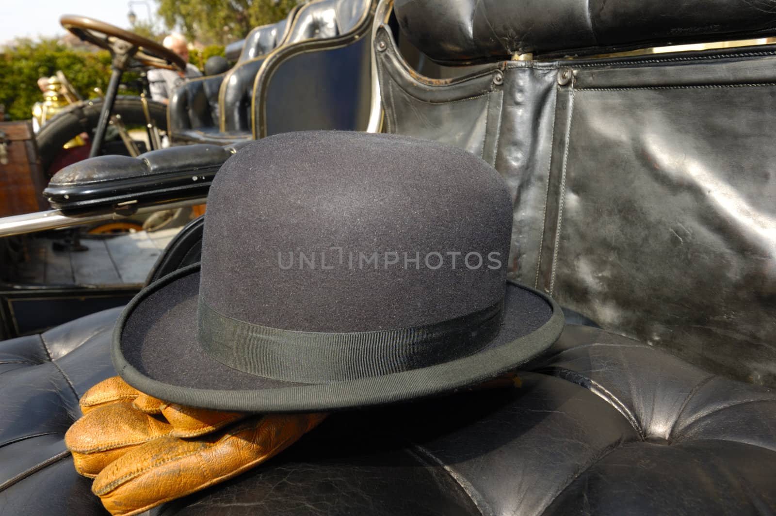 The black leather seat of a vintage motor-car with the driver's bowler hat and brown leather gloves placed carefully on it. Other vintage cars, lined up, can be seen in the background.