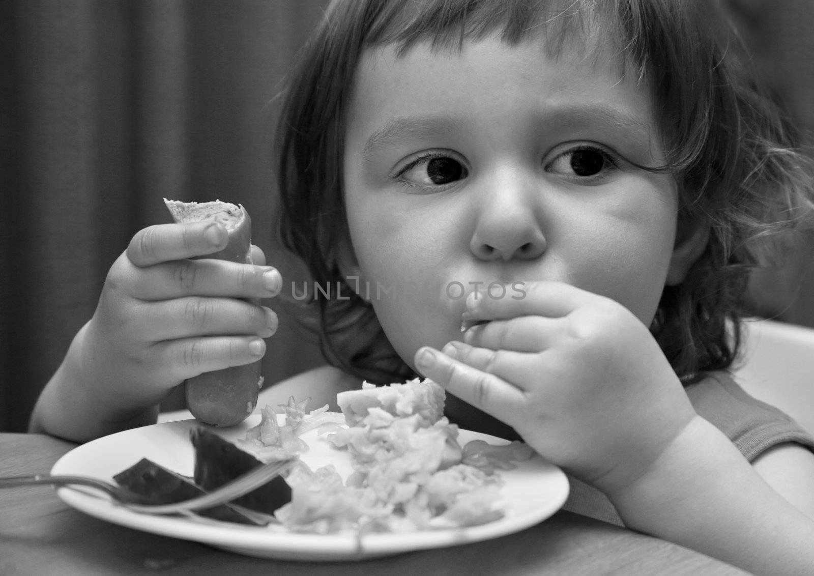 The small child eats sausage with macaroni. b/w