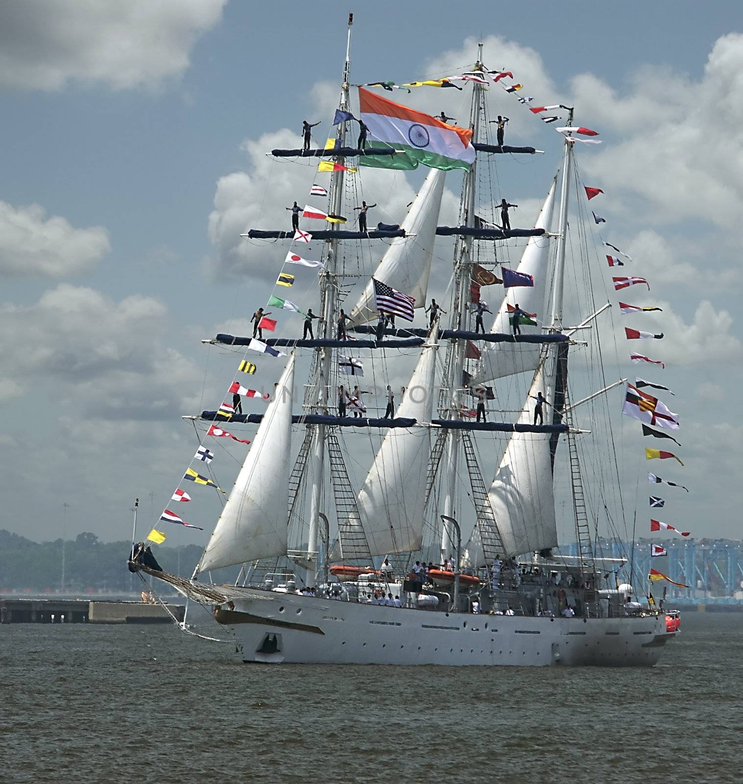 Sailors man the rails and masts of a sailing ship as it cruises into a coastal city's port