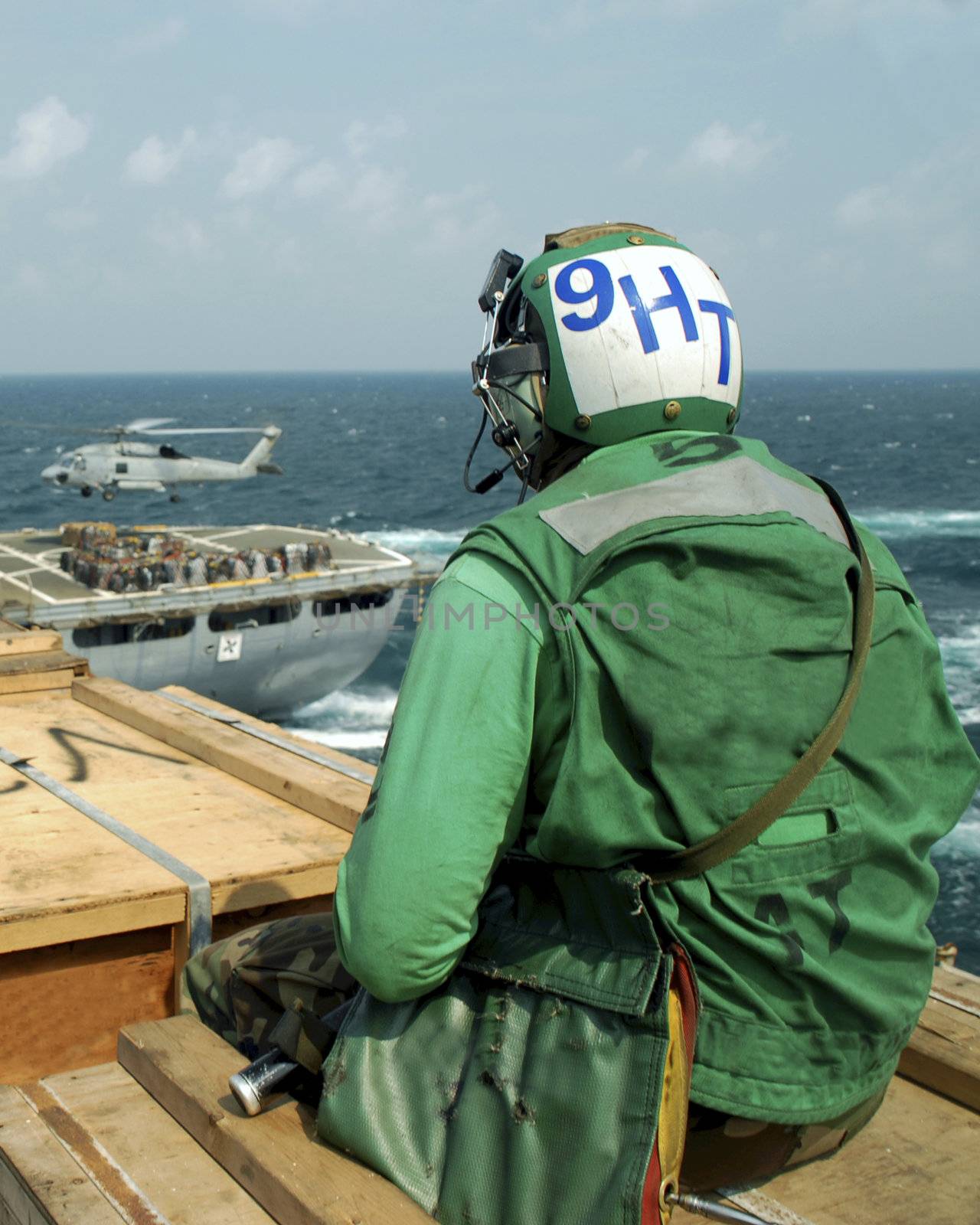 A sailor sits and watches a helicopter operate on a nearby ship