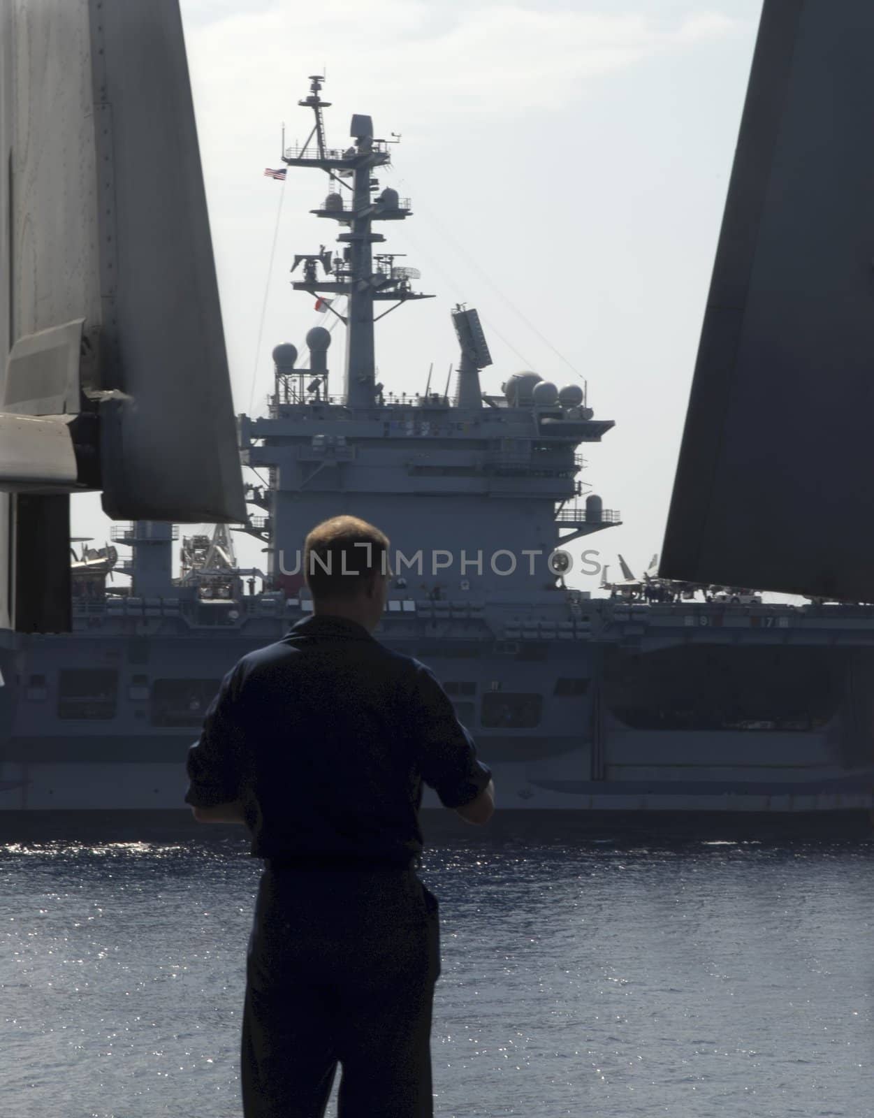 A sailor watches longingly as an aircraft carrier passes his ship on its way home
