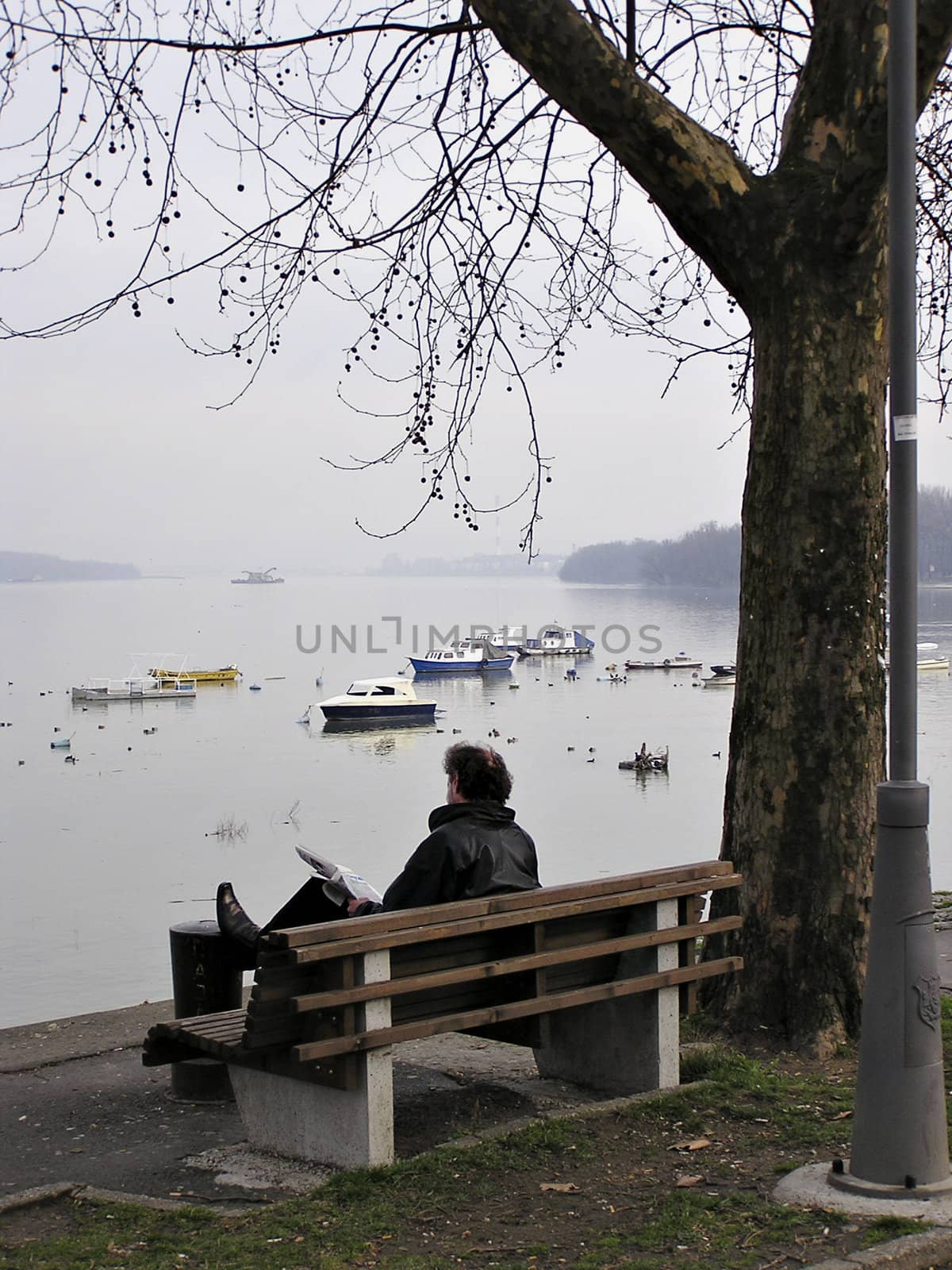Man sitting on bench at river bank reading newspapers.