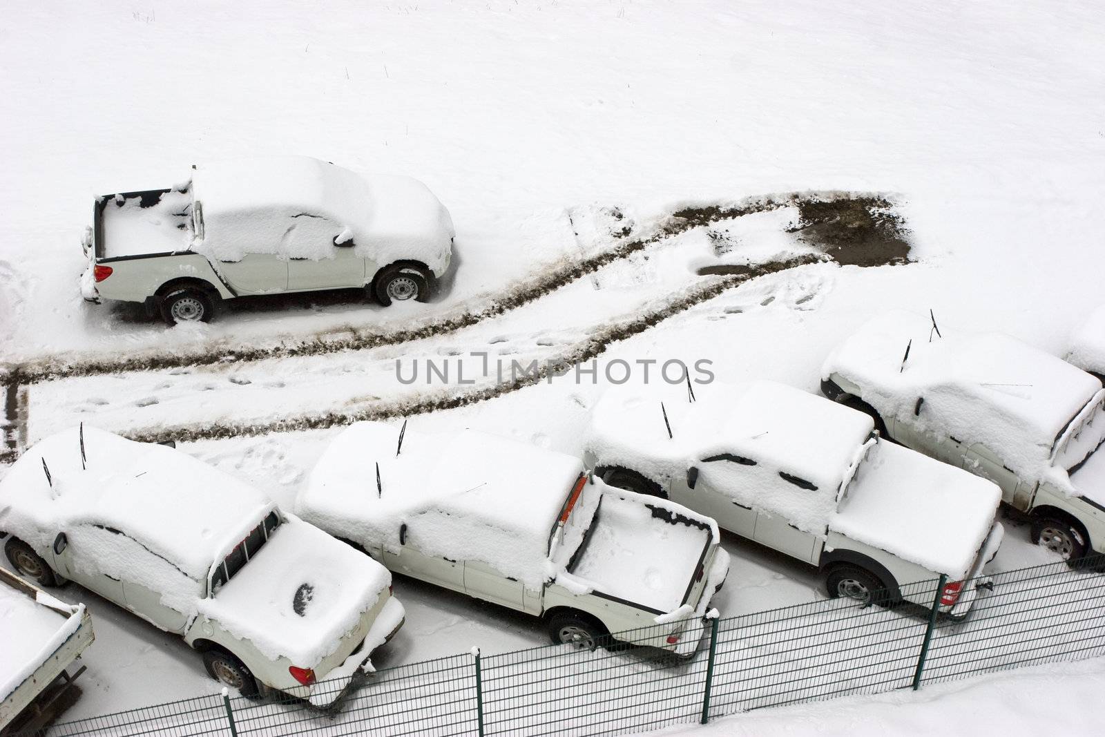 Parked cars under snow at parling lot with tracks of one gone car aerial view