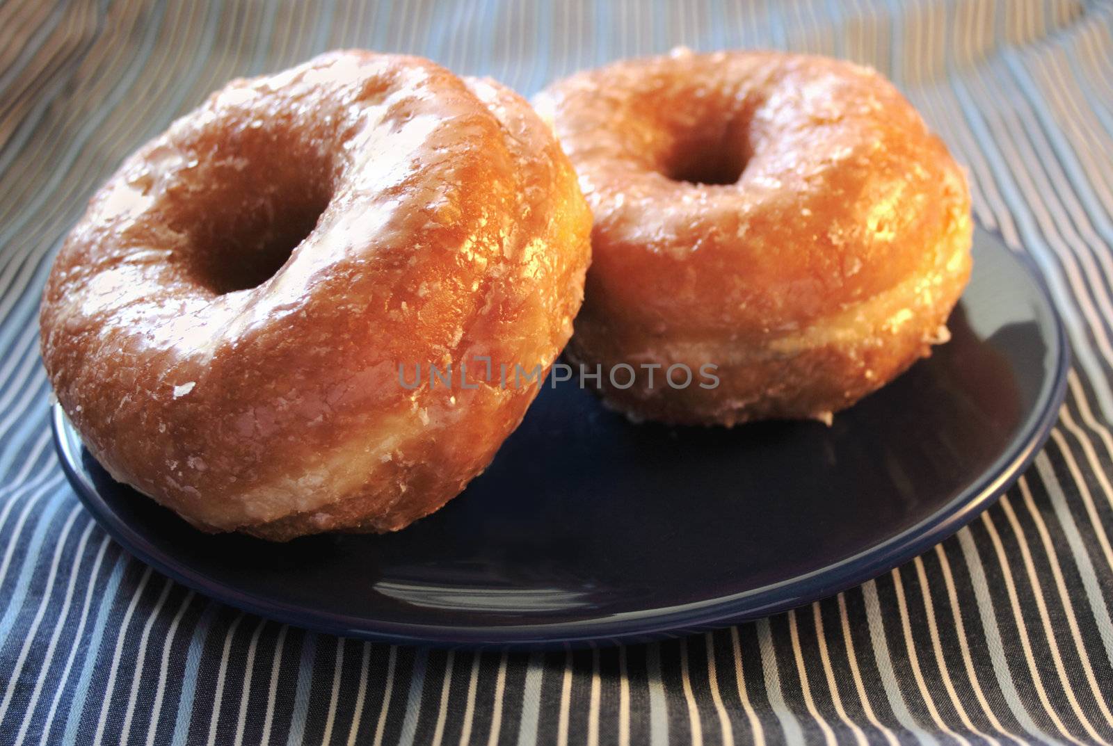 two glazed yeast doughnuts on a blue plate on a striped cloth
