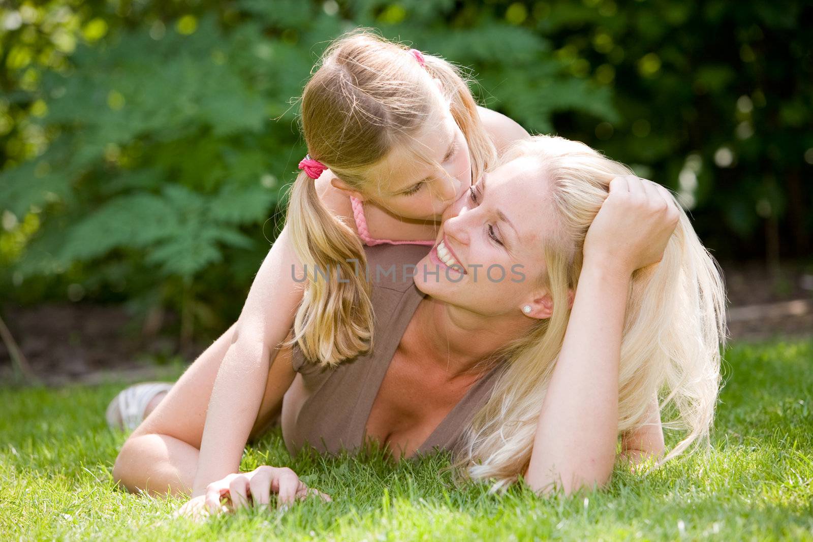 Pretty blond woman lying in the grass with her daughter, who is giving mum a kiss