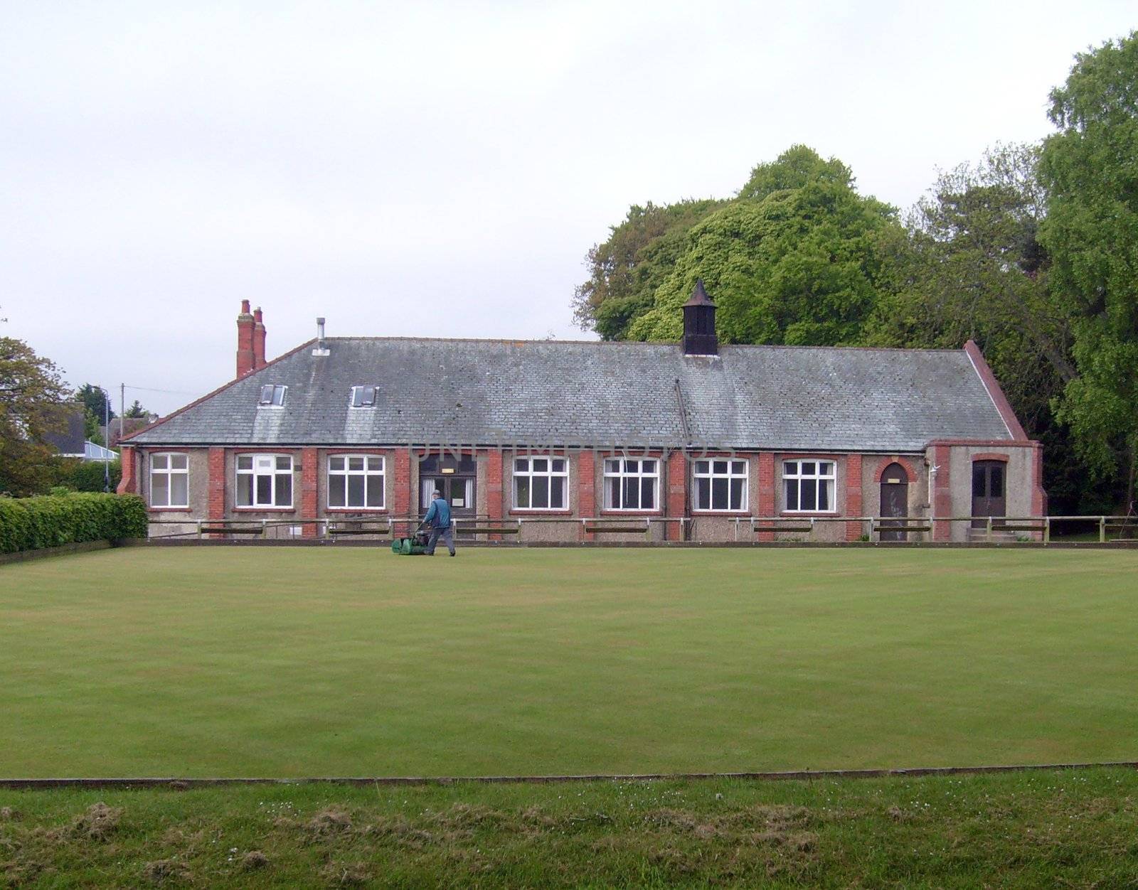 English village bowling green and pavilion on summers day, with groundsman cutting grass.