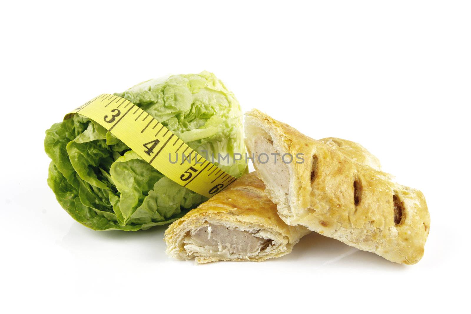 Contradiction between healthy food and junk food using a green salad lettace and sausage roll with a yellow tape measure on a reflective white background 