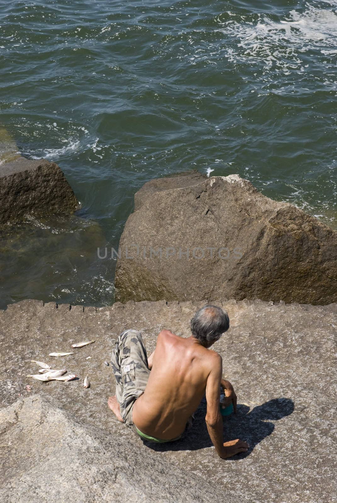High angle rear shot of an old man fishing on top of rocks, with sea directly below.