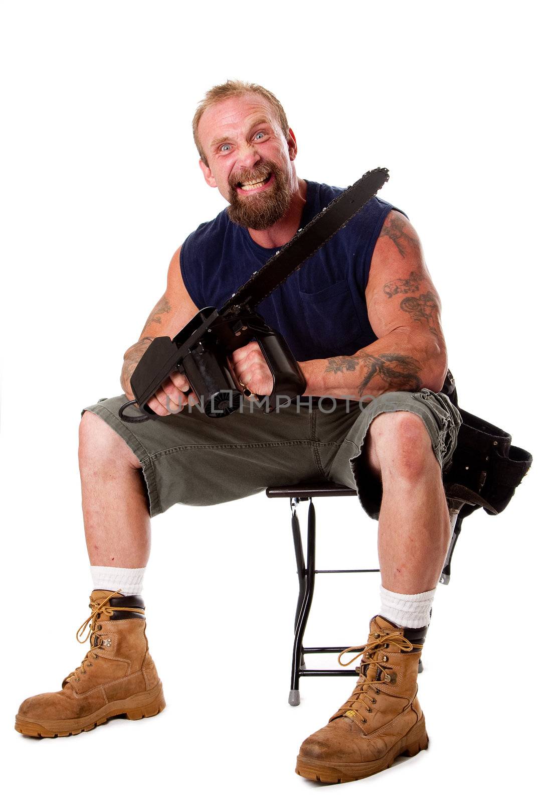 Crazy Caucasian man with tattoos and chainsaw sitting on stool with strong facial expression, isolated.