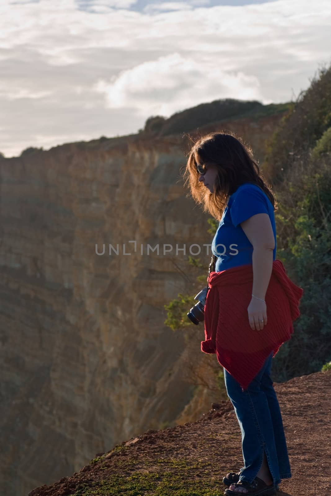A woman with a camera at the edge of a cliff, looking out, with more cliffs in the background.  Near Lagos, Portugal.