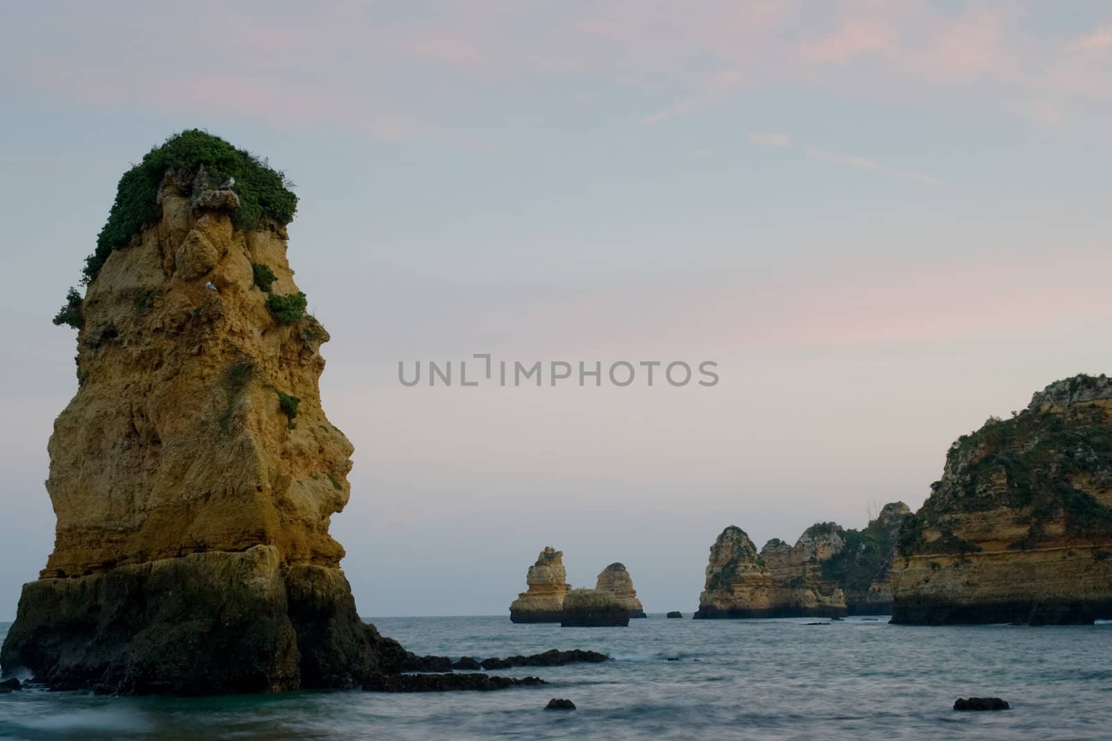 Along the beaches in Lagos, Portugal, a number of sheer rocky outcrops, as the sun sets casting pinks and blues in the sky.