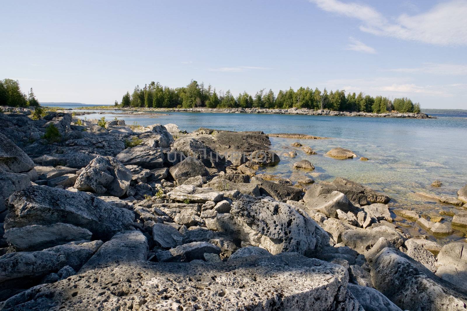 A rocky beach, clear water, clear day, and view of a small island in the distance.