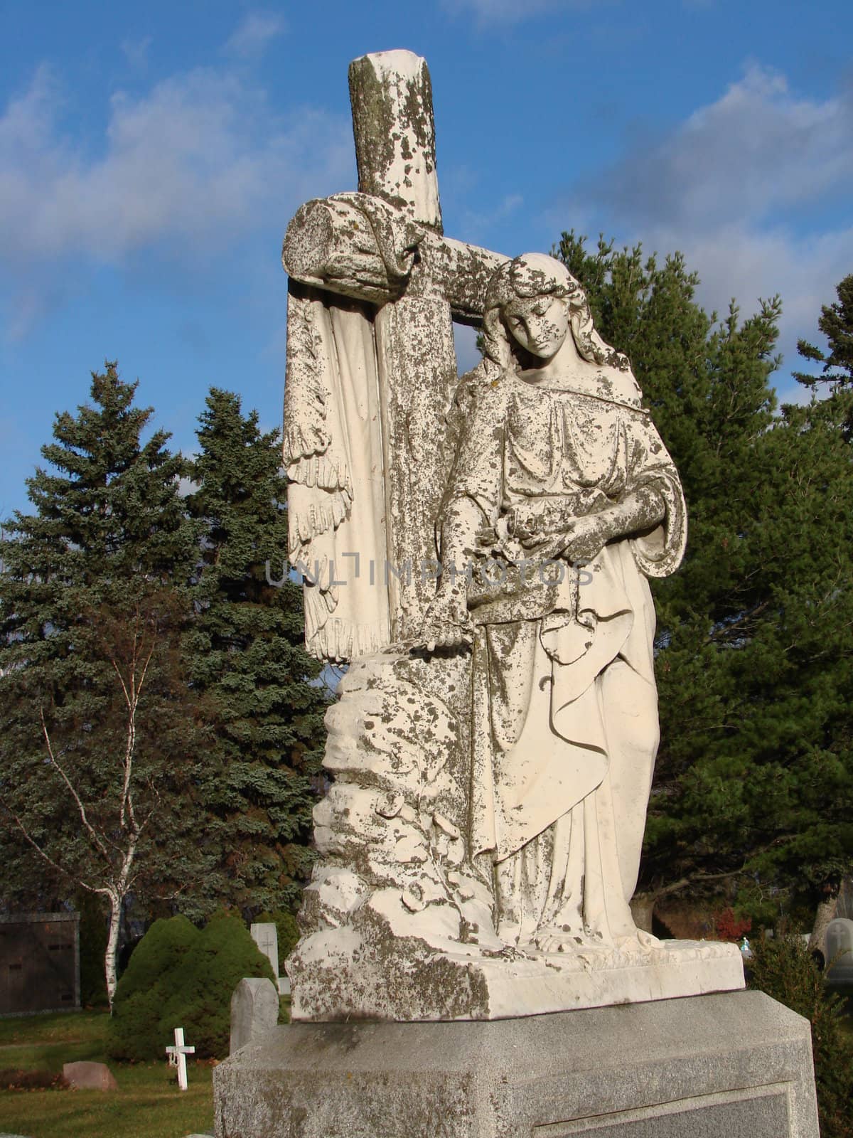White marble statue of Mary in Catholic Cemetery. Statue has moss and lichen on it. Fall day with bright blue sky and clouds.