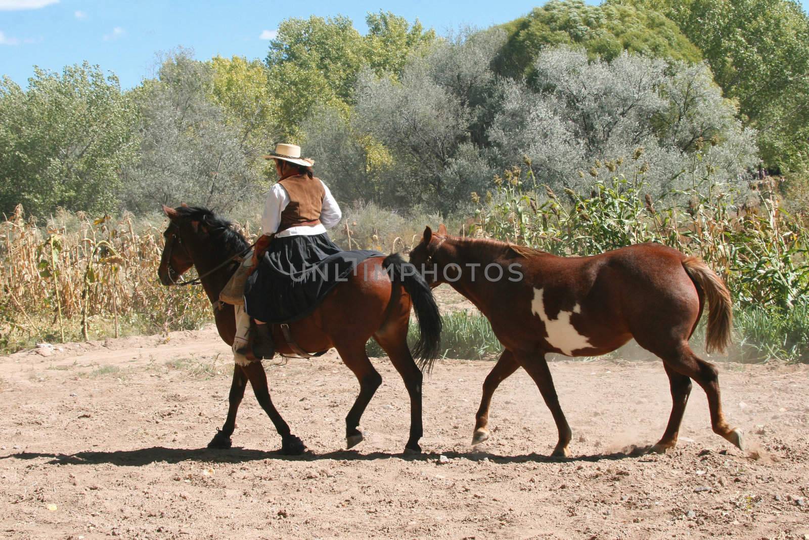 Cowgirl dressed in traditional attire leads paint horse down dusty road in New Mexico