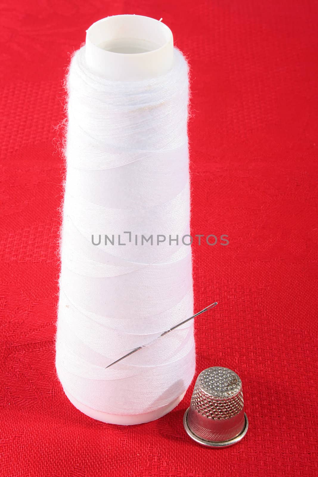 The coil with white threads and a needle on a red background, nearby a thimble for sewing.