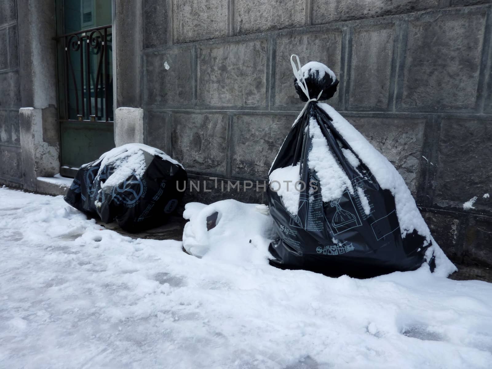 Garbage bags covered by snow in the street next to a building