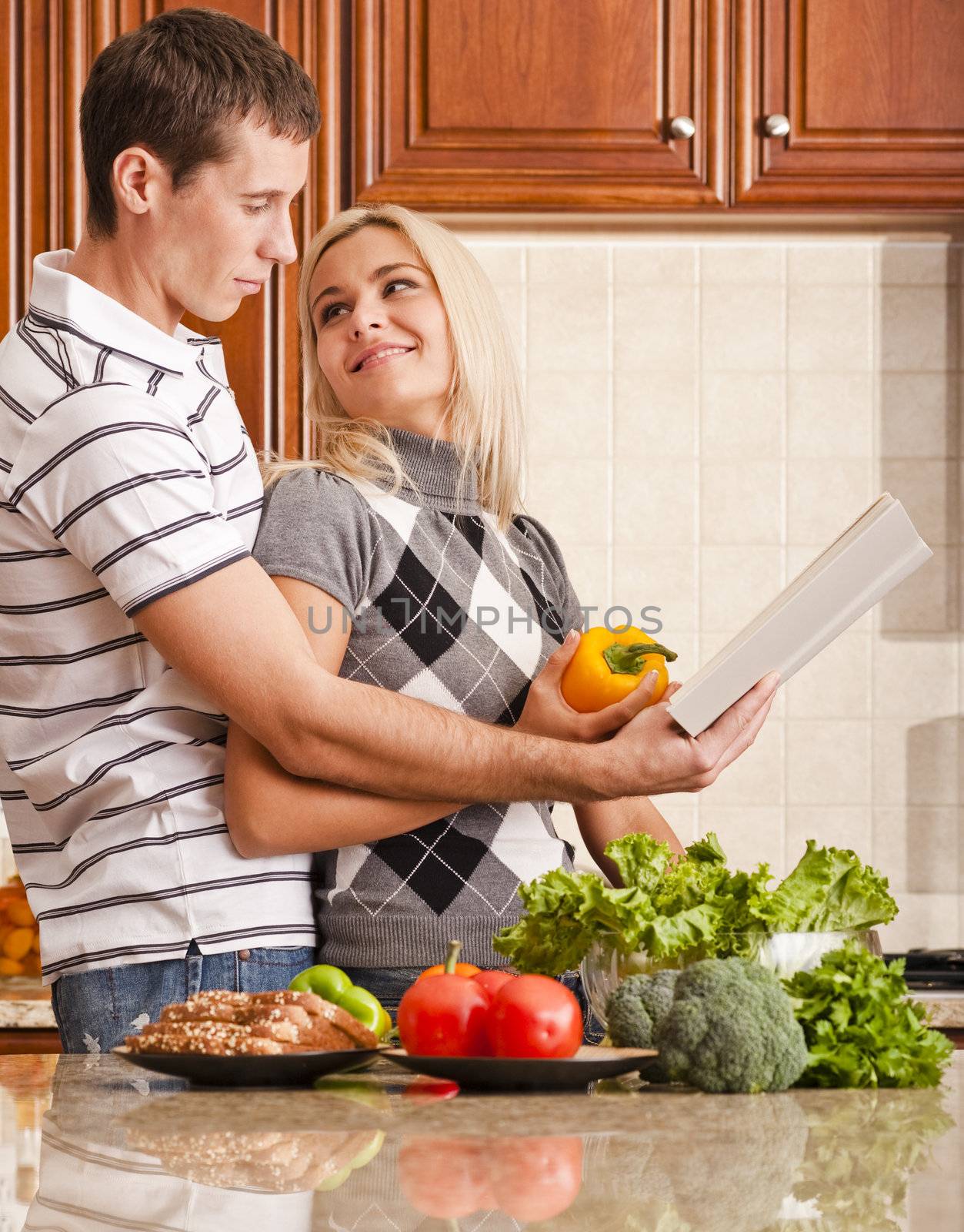 Young woman looks back affectionately at a young man as he reads a recipe book. A kitchen counter holds a variety of fresh vegetables in the foreground. Vertical shot.