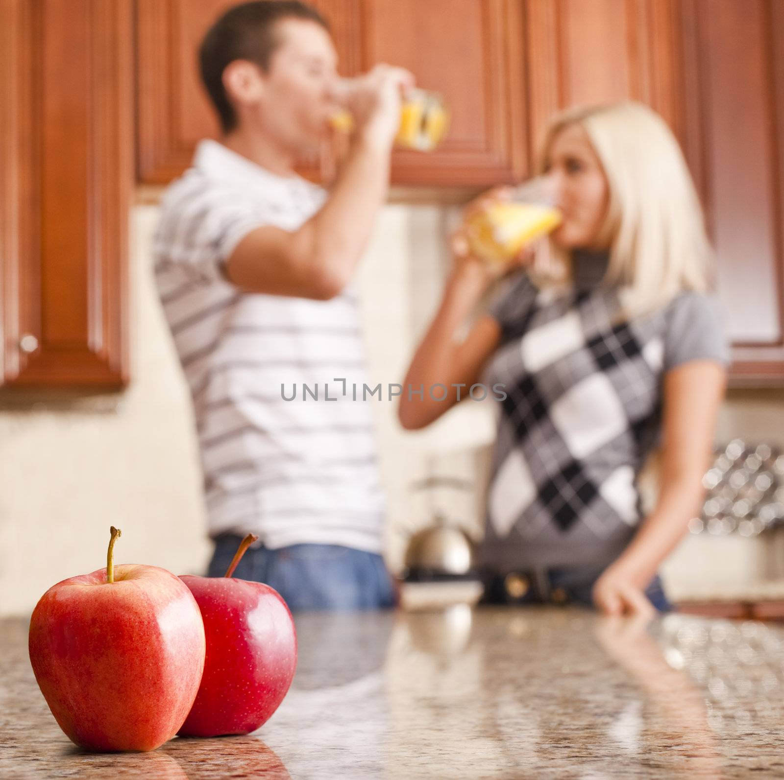 Young couple in kitchen drinking a glass of orange juice, two apples on counter in foreground. Square format.