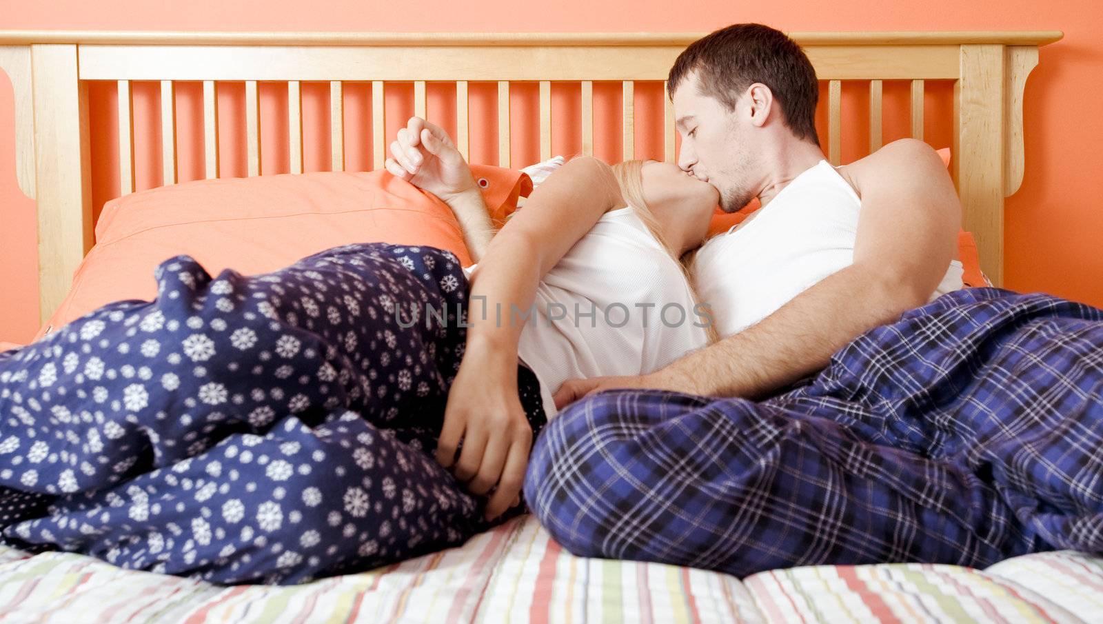 Young couple sitting on bed with stripped bedspread kissing. Horizontal shot.