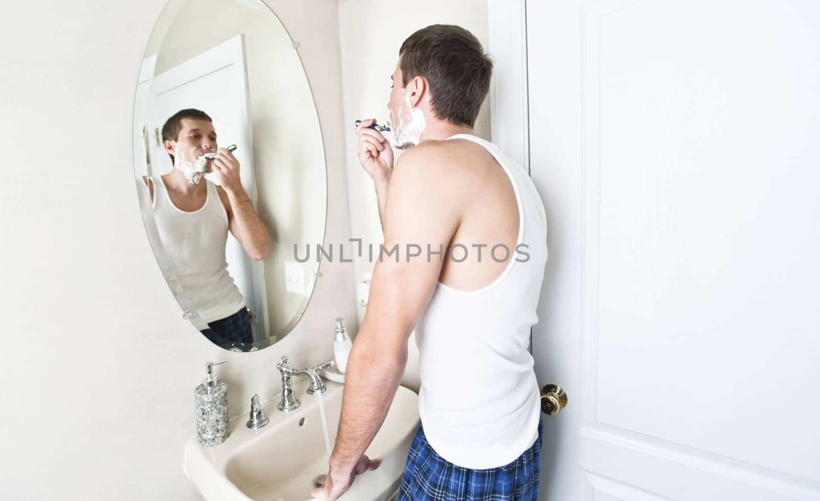 Young man in bathroom looking in the mirror and shaving. Horizontal shot.