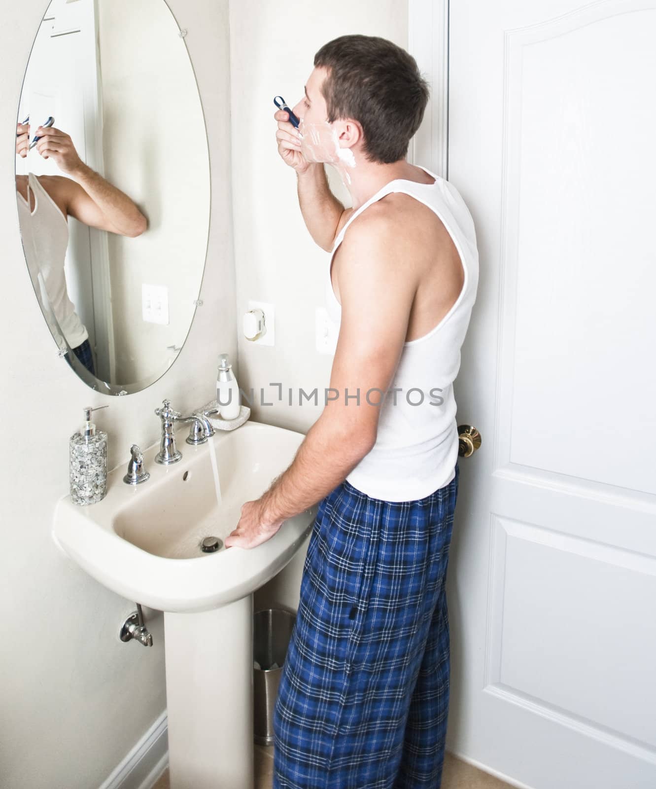 Young man in bathroom looking in the mirror and shaving. Vertical shot.