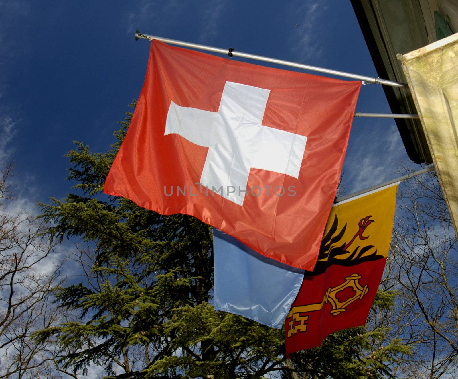 The flag of the Swiss Confederation with the flag of the canton of Geneva behind, against a blue sky laced with fine clouds.