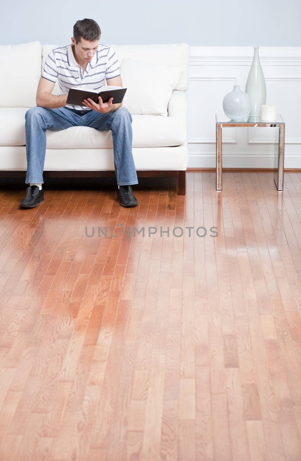 Young man in striped shirt and blue jeans sitting on white sofa reading a book. Vertical shot.