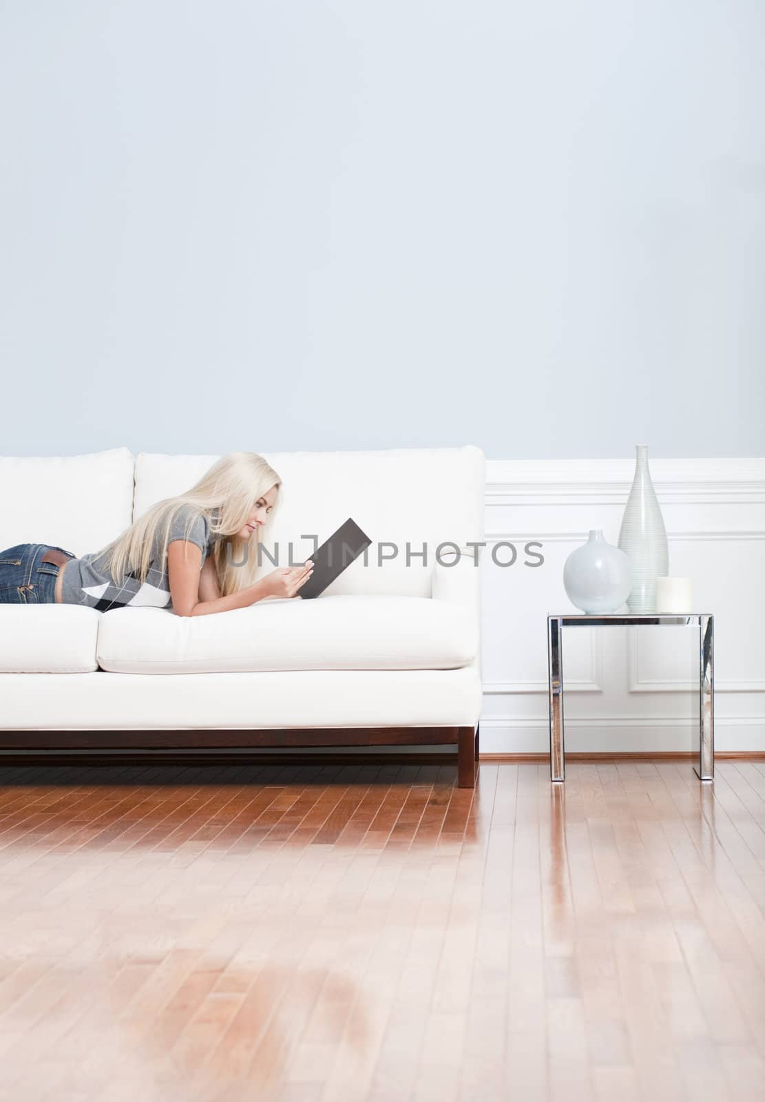 Young woman lies on a white sofa in a checkered top and blue jeans while reading a book. Vertical shot.