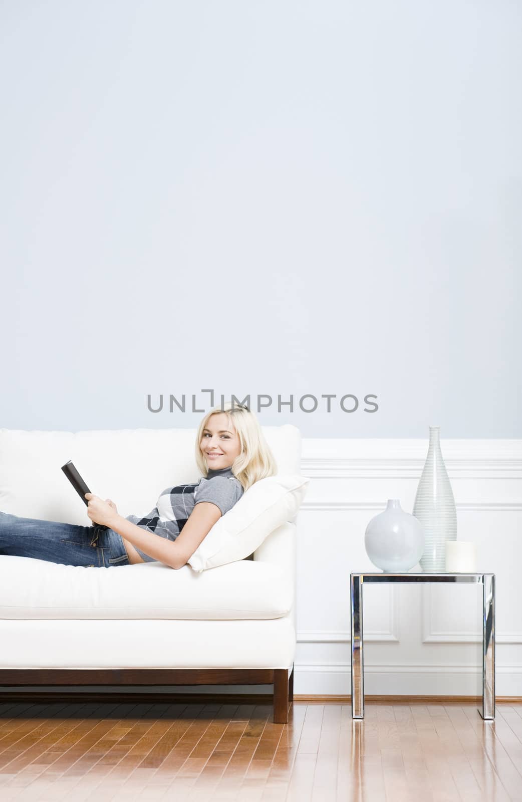 Young woman wears a checkered top and blue jeans lies on a white sofa while reading a book. Vertical shot.