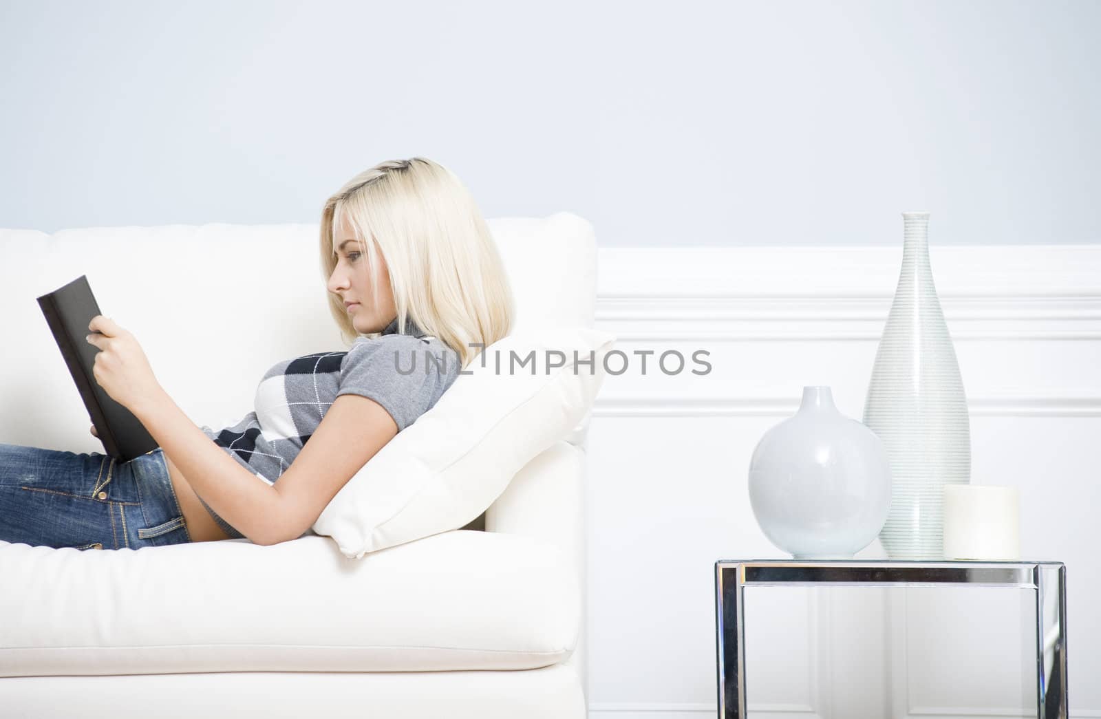 Young woman wears a checkered top and blue jeans, lying on white sofa and reading book. Hoizontal shot.