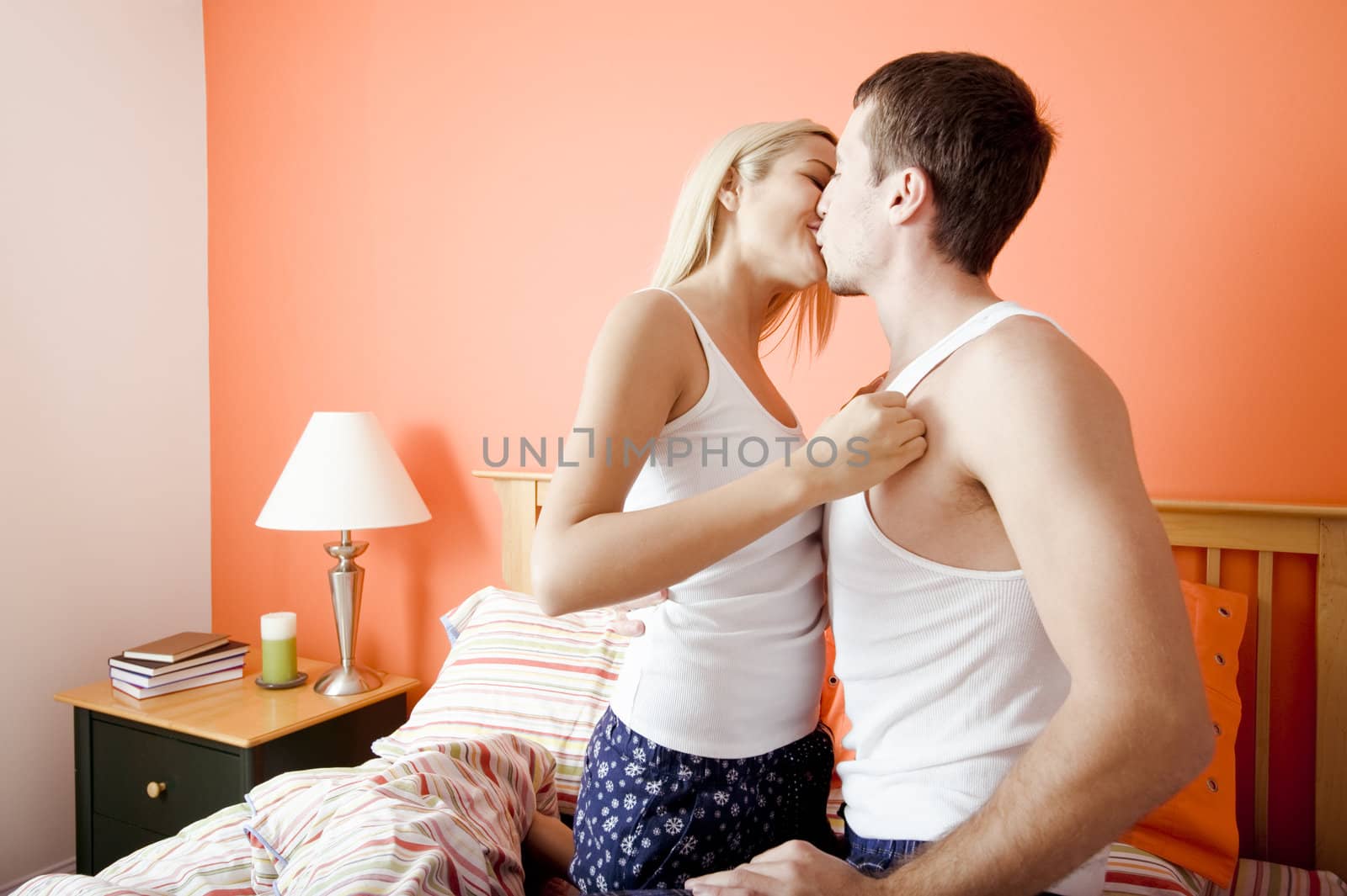 Young couple wearing white tank tops kiss passionately while kneeling on bed. Horizontal shot.